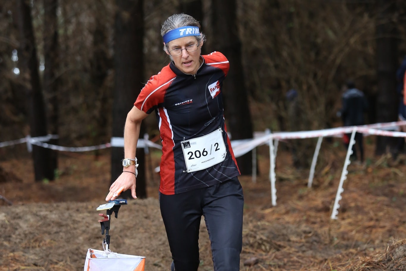 Christina Freeman punching a control in pine forest at the 2025 New Zealand Orienteering Championships relay