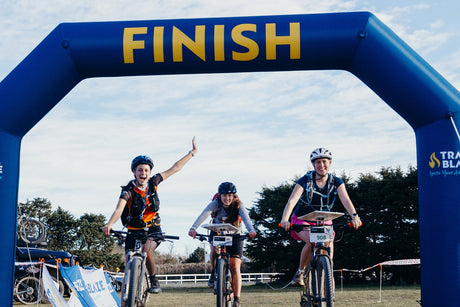 Three adventure racers riding mountain bikes across the TrailBlaze finish arch, one with an arm raised